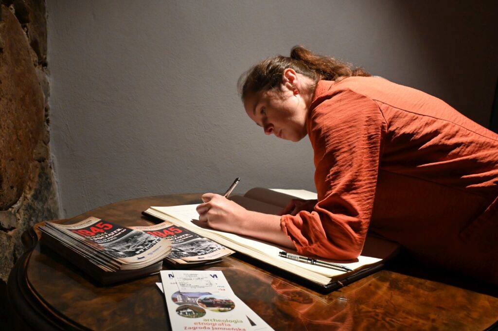 Członkini francuskiej delegacji wpisuje się do księgi pamiątkowej muzeum przy drewnianym stoliku, obok leżą materiały promocyjne (kliknięcie powiększy zdjęcie).