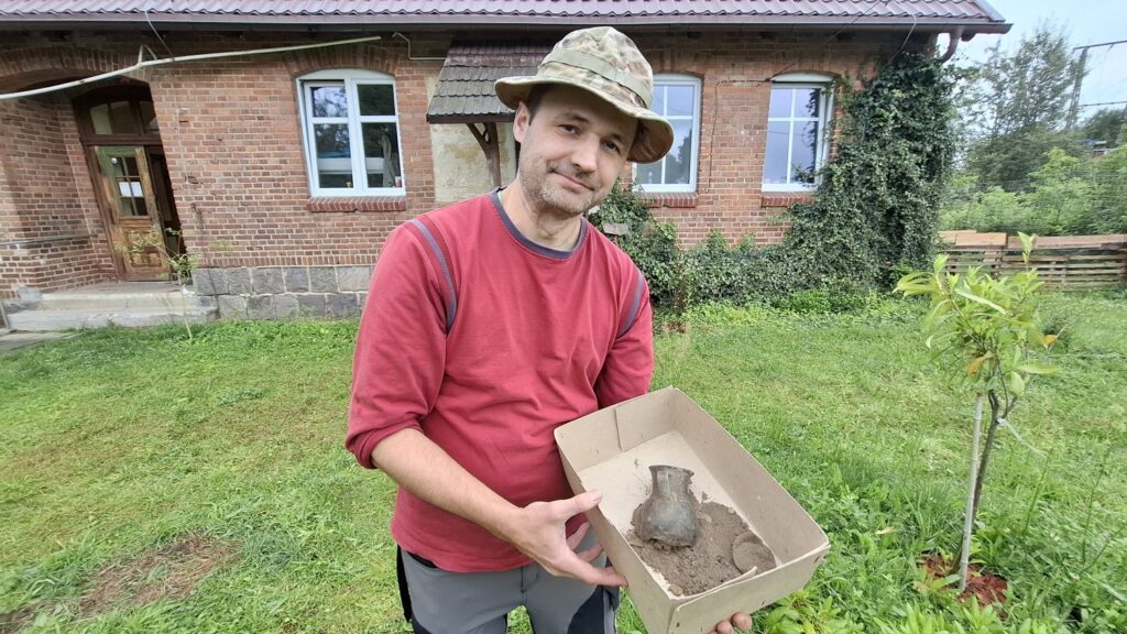 Archeolog prezentuje, ułożony w kartonowym pudle, fragmenty dzbanka. W tle ceglane zabudowania (kliknięcie spowoduje powiększenie zdjęcia).
