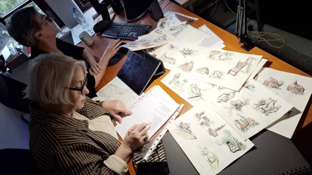 Dwie kobiety siedzą przed biurkiem, na którym rozłożone są projekty rysunków różnych obiektów architektonicznych (kliknięcie spowoduje powiększenie zdjęcia).