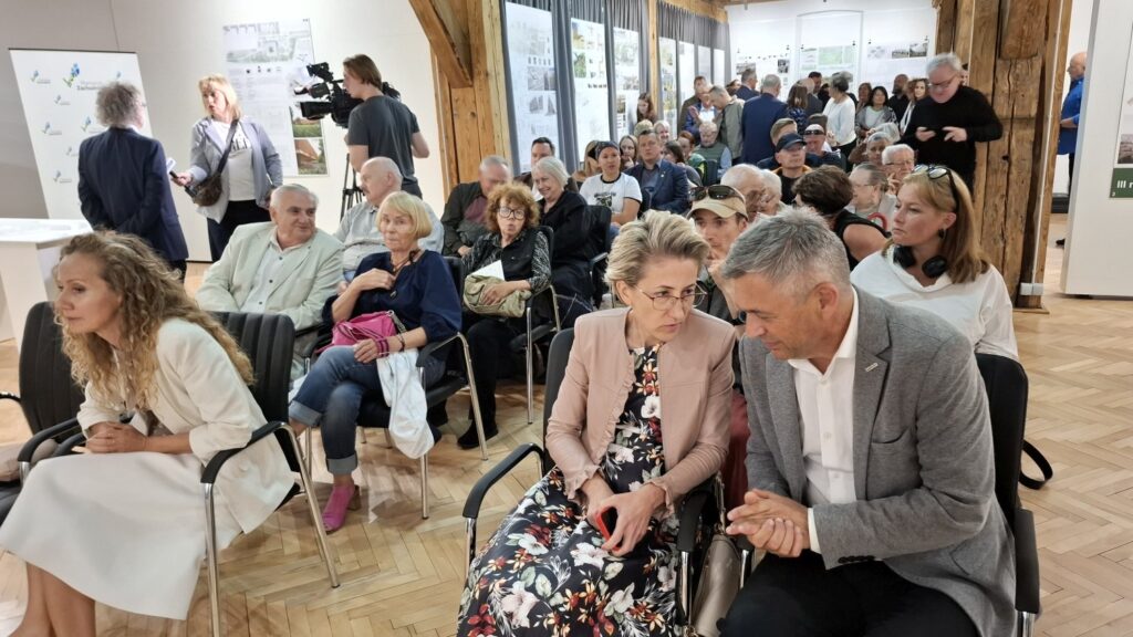 Publiczność zgromadzona w sali wystawowej, uczestnicy siedzą na krzesłach i rozmawiają przed rozpoczęciem wydarzenia (kliknięcie powiększy zdjęcie).