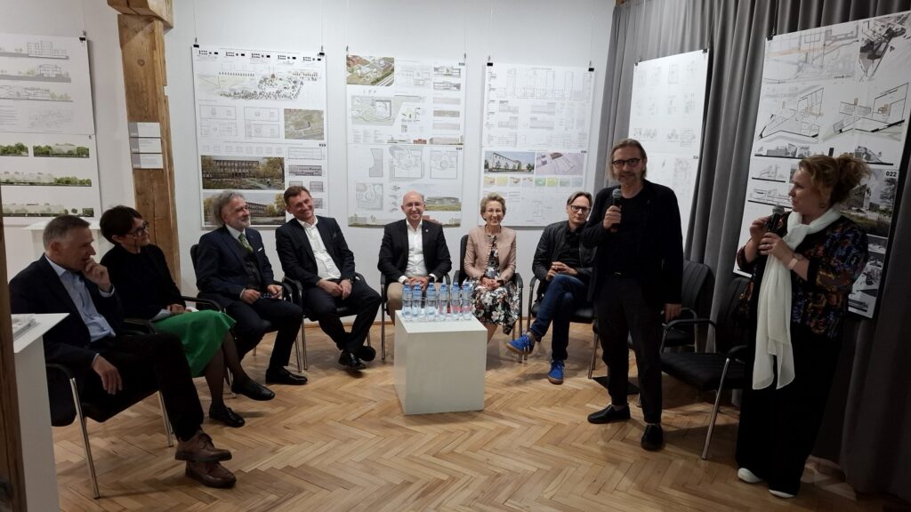 Panel dyskusyjny z udziałem kilku osób siedzących przy stole, jedna z kobiet stoi i przemawia do publiczności (kliknięcie powiększy zdjęcie).