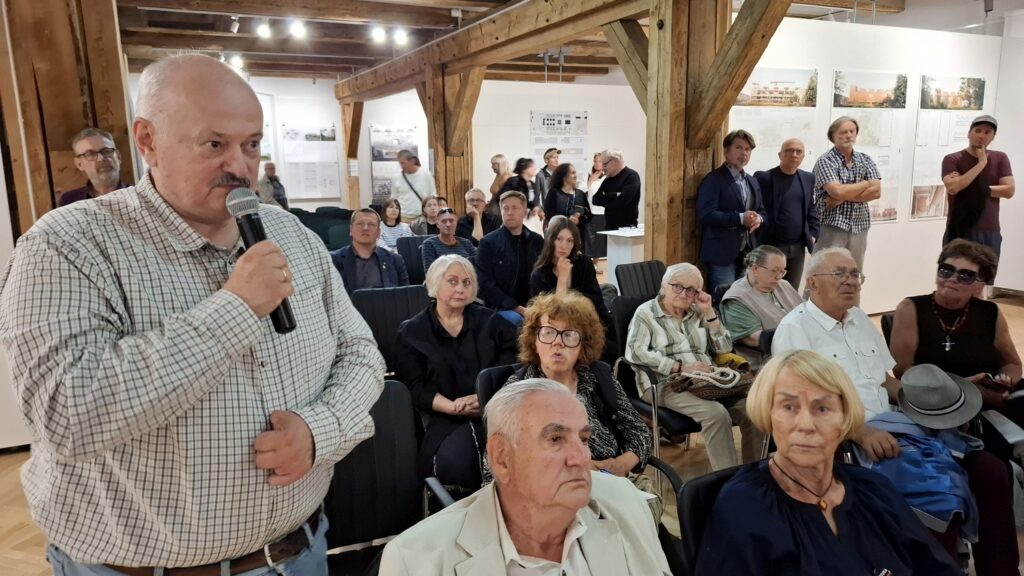 Mężczyzna trzymający mikrofon zadaje pytanie podczas spotkania, w tle siedzą uczestnicy wydarzenia (kliknięcie powiększy zdjęcie).