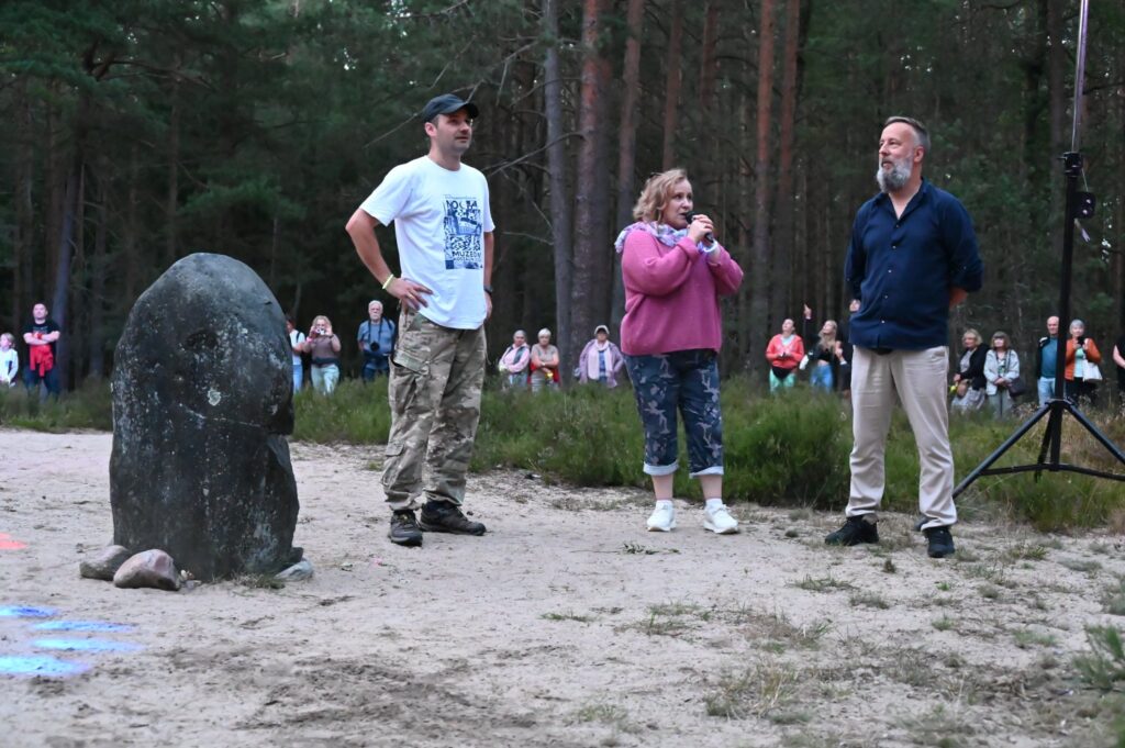 Organizatorzy przemawiają do uczestników Nocy Gotów przy kamieniu w lesie, a zgromadzona publiczność słucha w tle (kliknięcie powiększy zdjęcie).
