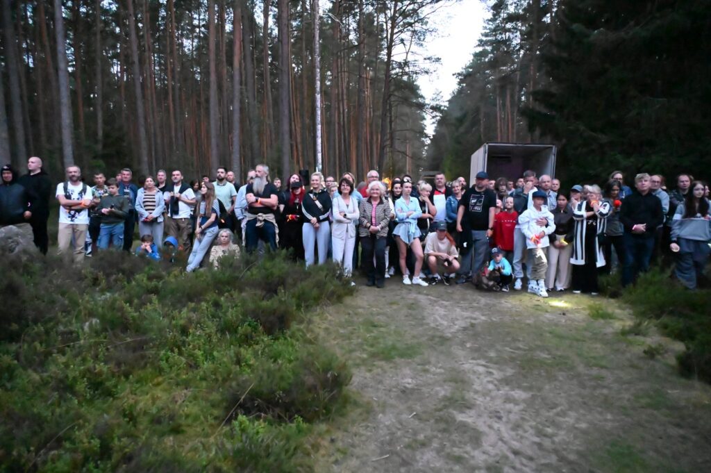Liczna grupa uczestników gromadzi się na leśnej ścieżce podczas wydarzenia, stojąc wśród wysokich drzew (kliknięcie powiększy zdjęcie).