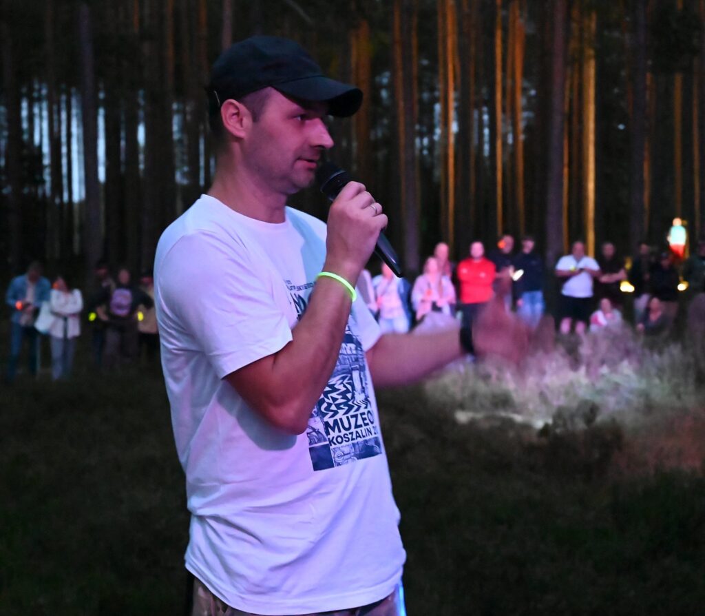 Andrzej Kasprzak przemawia do zgromadzonych uczestników w lesie, trzymając mikrofon, a publiczność stoi w tle i słucha (kliknięcie powiększy zdjęcie).