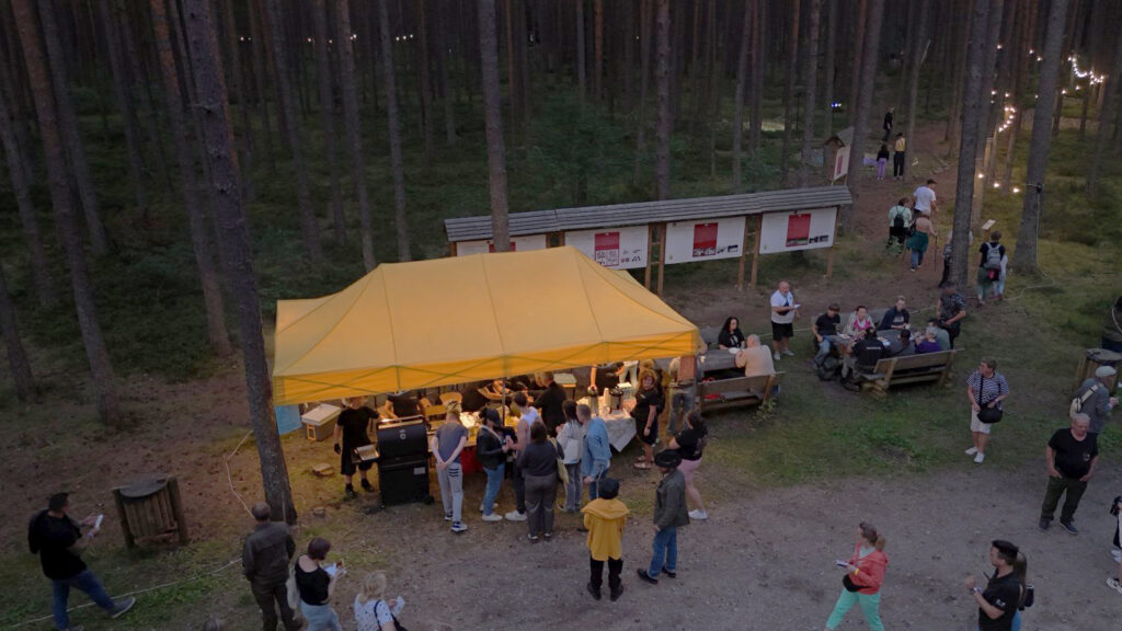 Widok z drona na wydarzenie w lesie – grupa osób gromadzi się wokół namiotu i stołów wśród drzew (kliknięcie powiększy zdjęcie).