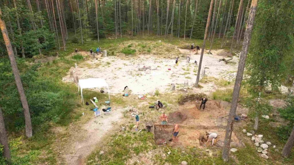 Szeroki widok z góry na teren wykopalisk w lesie z widocznym namiotem i pracującymi badaczami (kliknięcie powiększy zdjęcie).