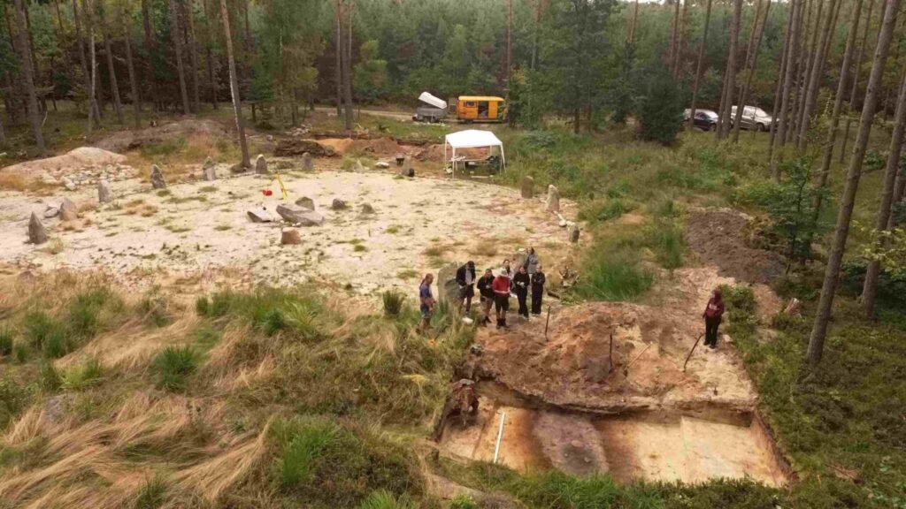 Widok z góry na stanowisko archeologiczne, w tle zaplecze techniczne i grupa osób przy wykopach (kliknięcie powiększy zdjęcie).