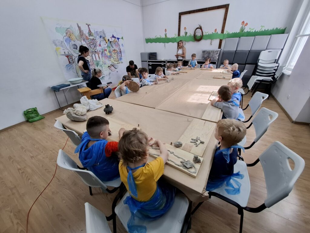 Dzieci siedzą przy stołach rozstawionych na sali edukacyjnej, obserwują prowadzącego zajęcia i podpatrują jak pracować na kole garncarskim (kliknięcie spowoduje powiększenie zdjęcia).