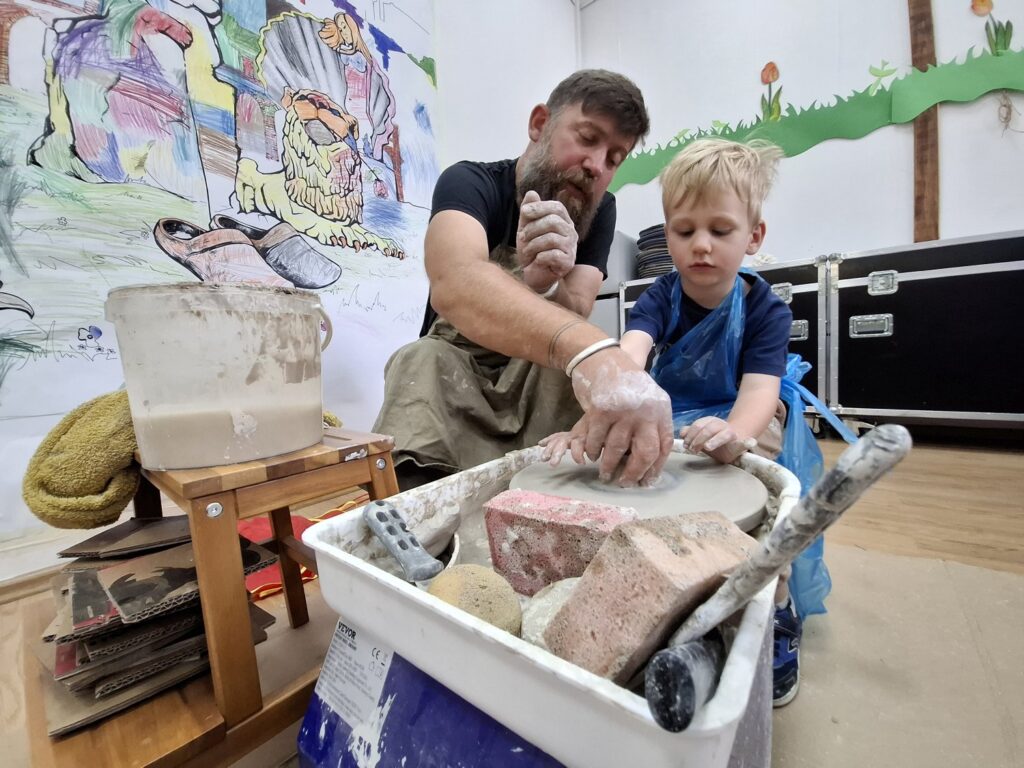 Chłopiec, w obecności dorosłego prowadzącego warsztaty, siedzi przy kole garncarskim i obrabia formę z gliny (kliknięcie spowoduje powiększenie zdjęcia).
