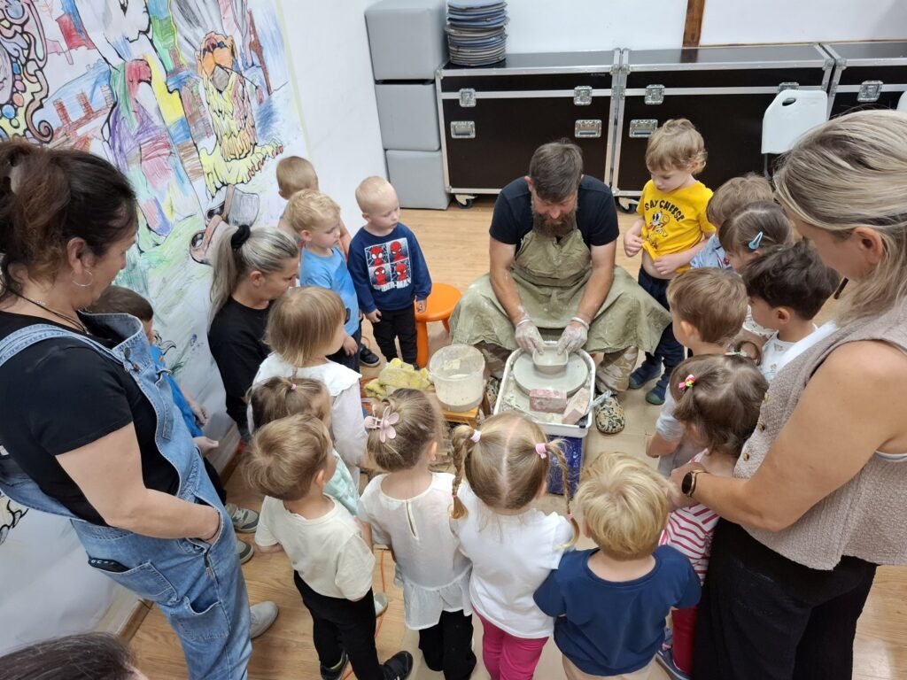 Dzieci stoją wokół osoby prowadzącej zajęcia i obserwują, jak pracuje się na kole garncarskim (kliknięcie spowoduje powiększenie zdjęcia).