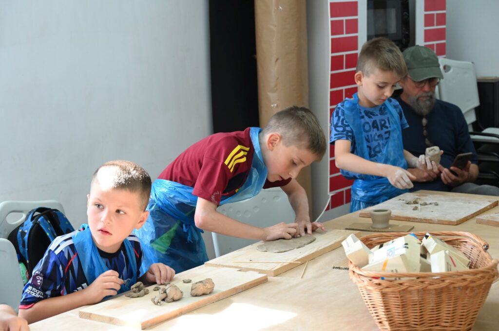 Dzieci siedzą przy stole i modelują przedmioty z gliny (kliknięcie spowoduje powiększenie zdjęcia).