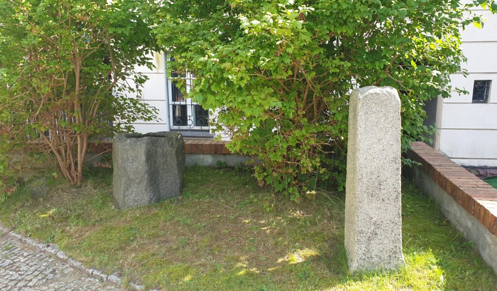 Średniowieczna chrzcielnica ustawiona na trawniku przy budynku muzeum; w tle widoczne niewielkie krzewy (kliknięcie powiększy zdjęcie).