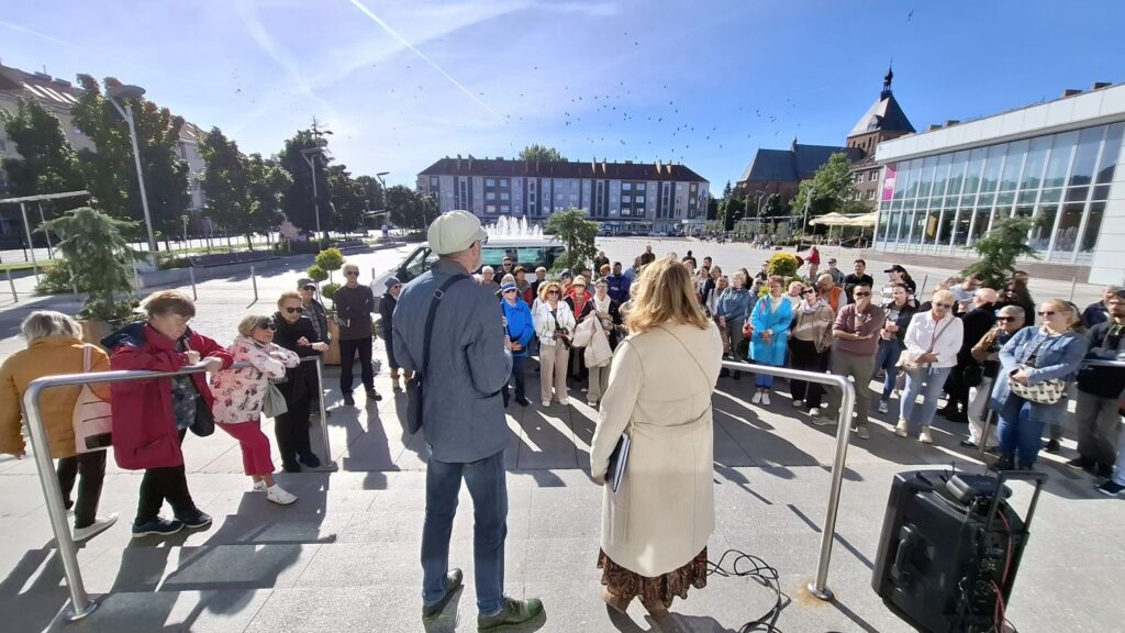 Przewodnik i uczestnicy spaceru historycznego stoją przed uczestnikami wydarzenia, w tle Rynek i zabudowania miasta oraz katedra (kliknięcie powiększy zdjęcie).