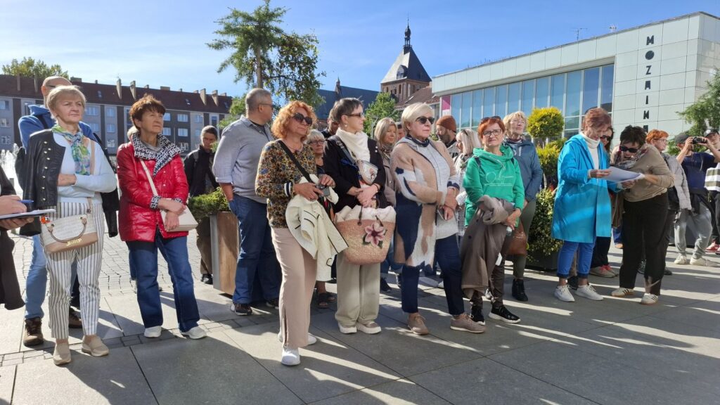 Zbliżenie na uczestników spaceru historycznego słuchających opowieści przewodnika (kliknięcie powiększy zdjęcie).