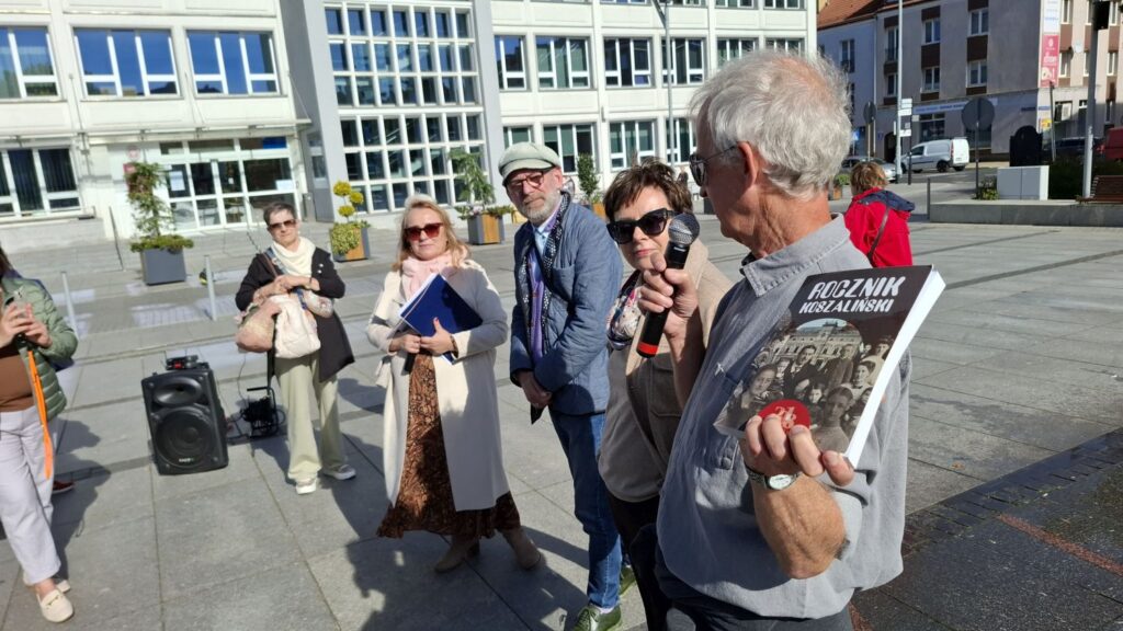 Lokalny historyk prezentuje publikację dotyczącą historii miasta, w tle pozostali uczestnicy wydarzenia oraz budynek Ratusza (kliknięcie powiększy zdjęcie).