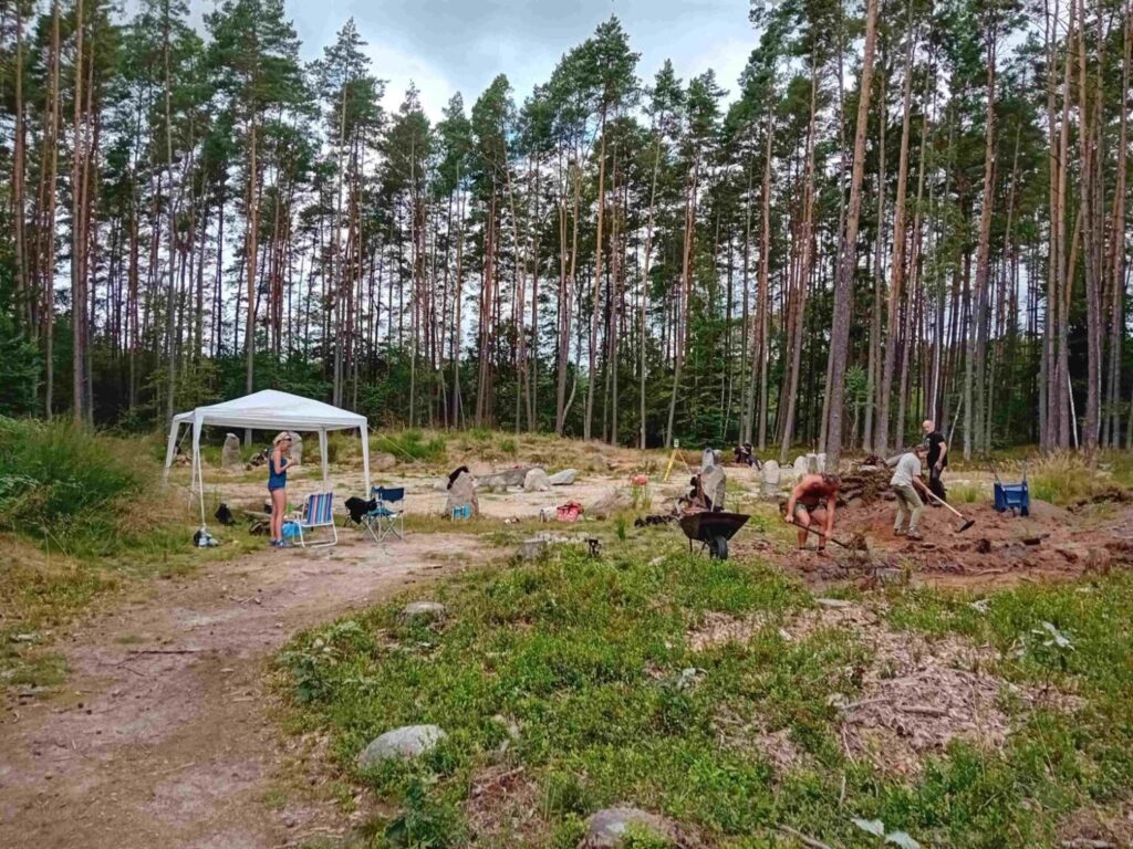 Stanowisko archeologiczne w lesie, widoczne wykopy oraz pracujący badacze i namiot techniczny (kliknięcie powiększy zdjęcie).