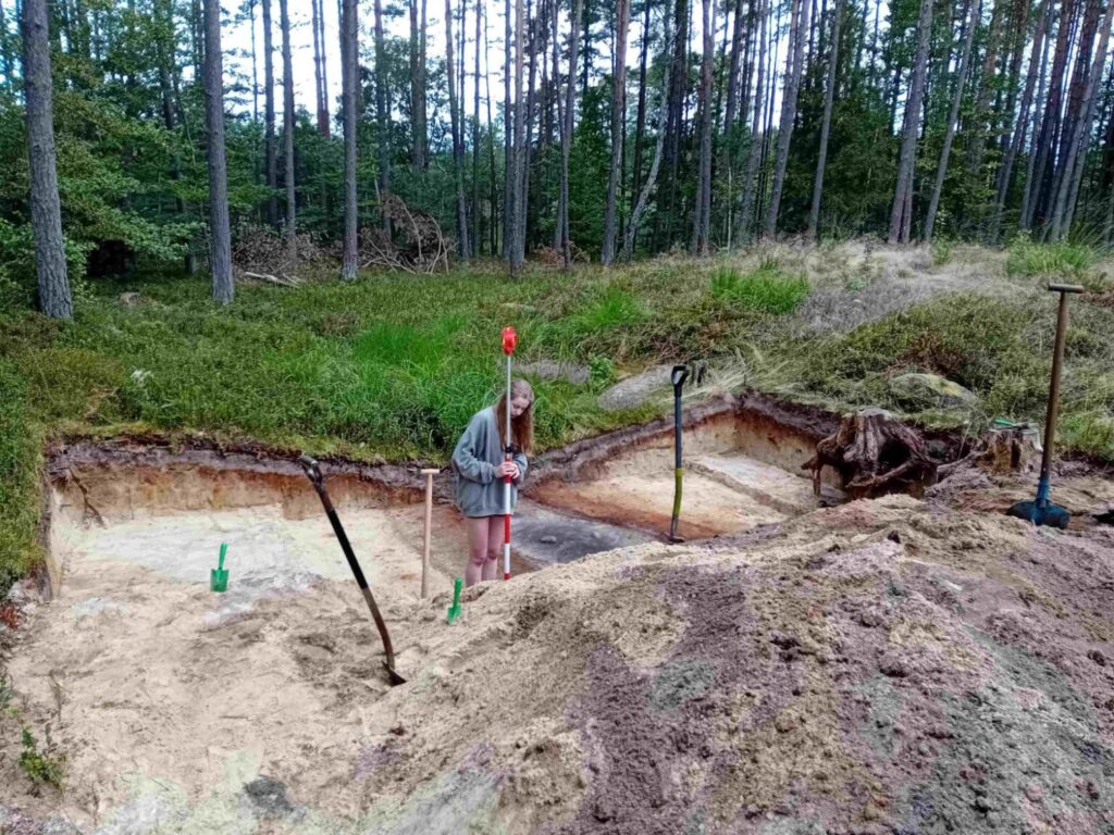 Osoba prowadzi prace pomiarowe na terenie wykopu archeologicznego wśród piaszczystych warstw gruntu (kliknięcie powiększy zdjęcie).