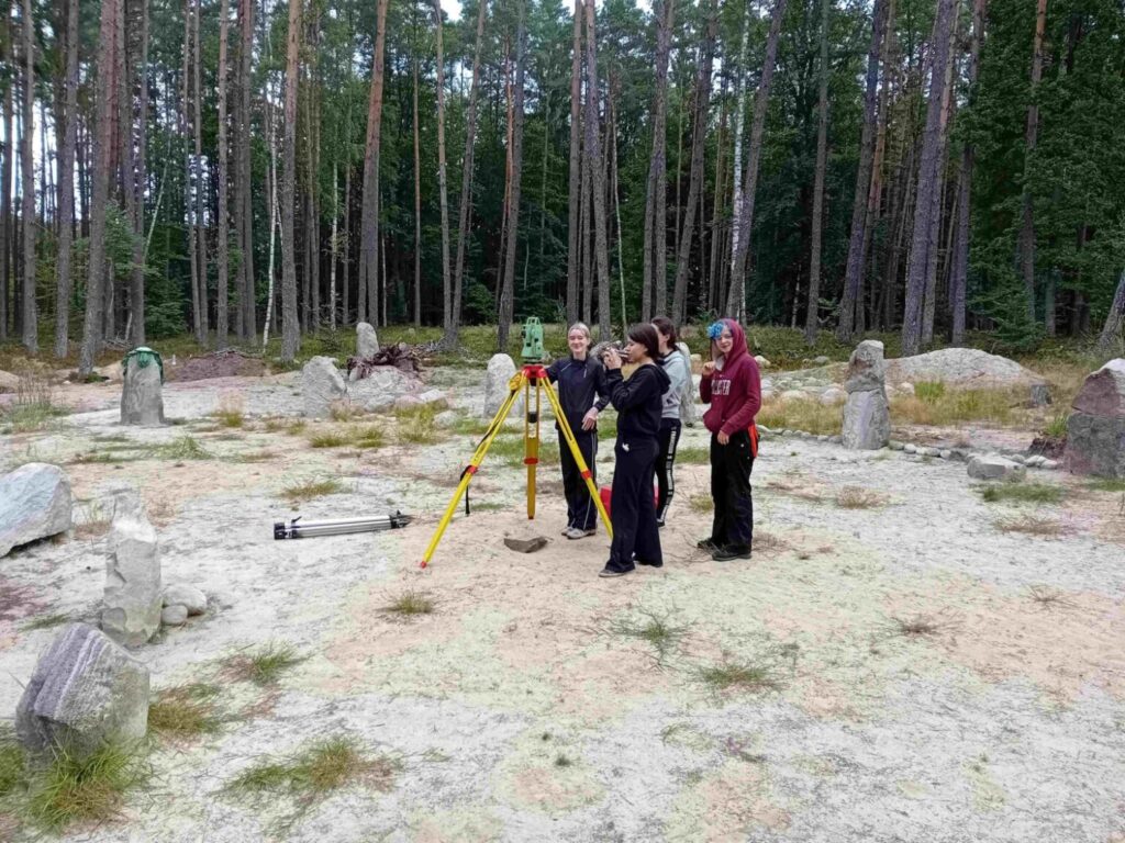 Grupa osób wykonuje pomiary geodezyjne na stanowisku archeologicznym w lesie (kliknięcie powiększy zdjęcie).