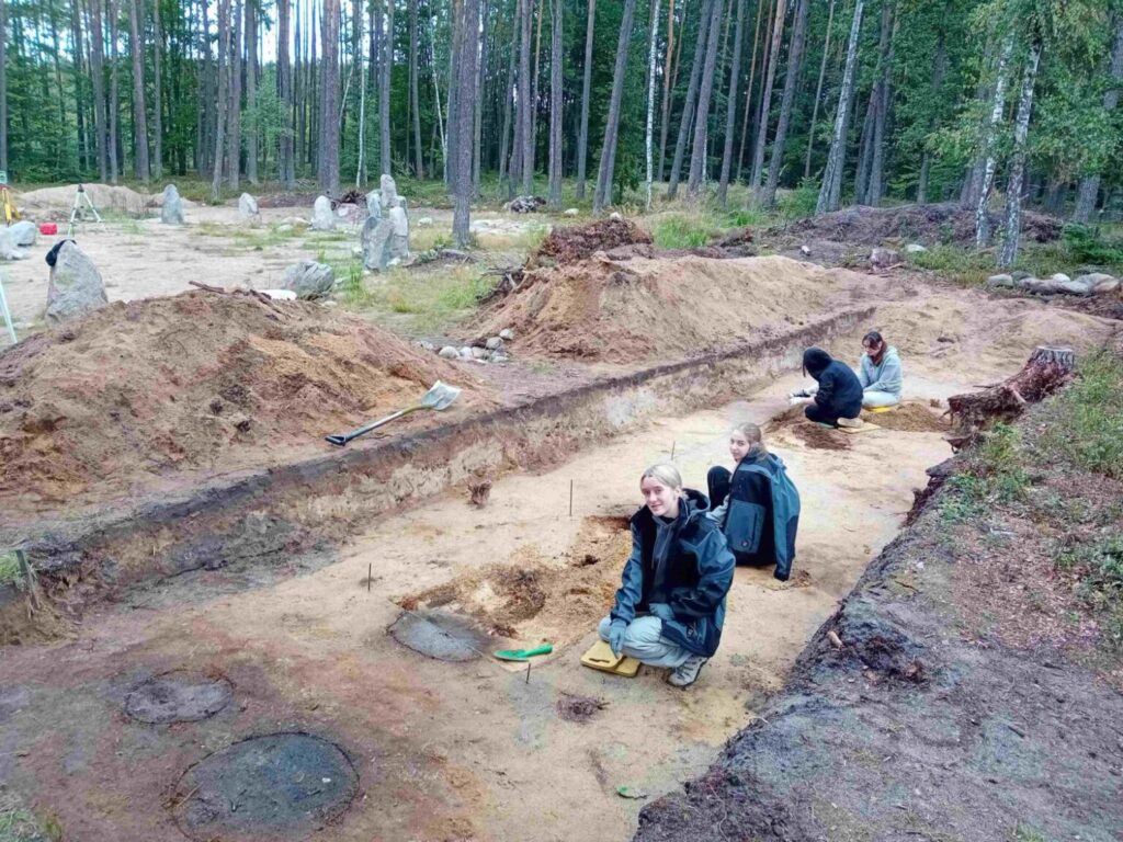 Archeolodzy prowadzą prace badawcze w wykopie, odsłaniając relikty w piaszczystym podłożu (kliknięcie powiększy zdjęcie).