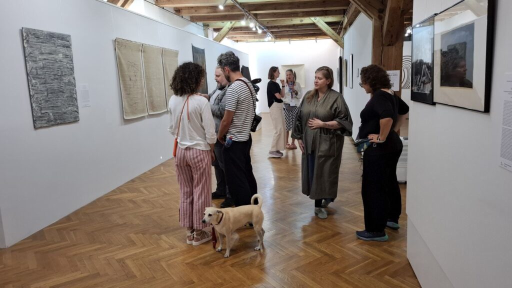 Zwiedzający oglądają prace na ścianach galerii, wśród nich stoi pies na smyczy (kliknięcie powiększy zdjęcie).