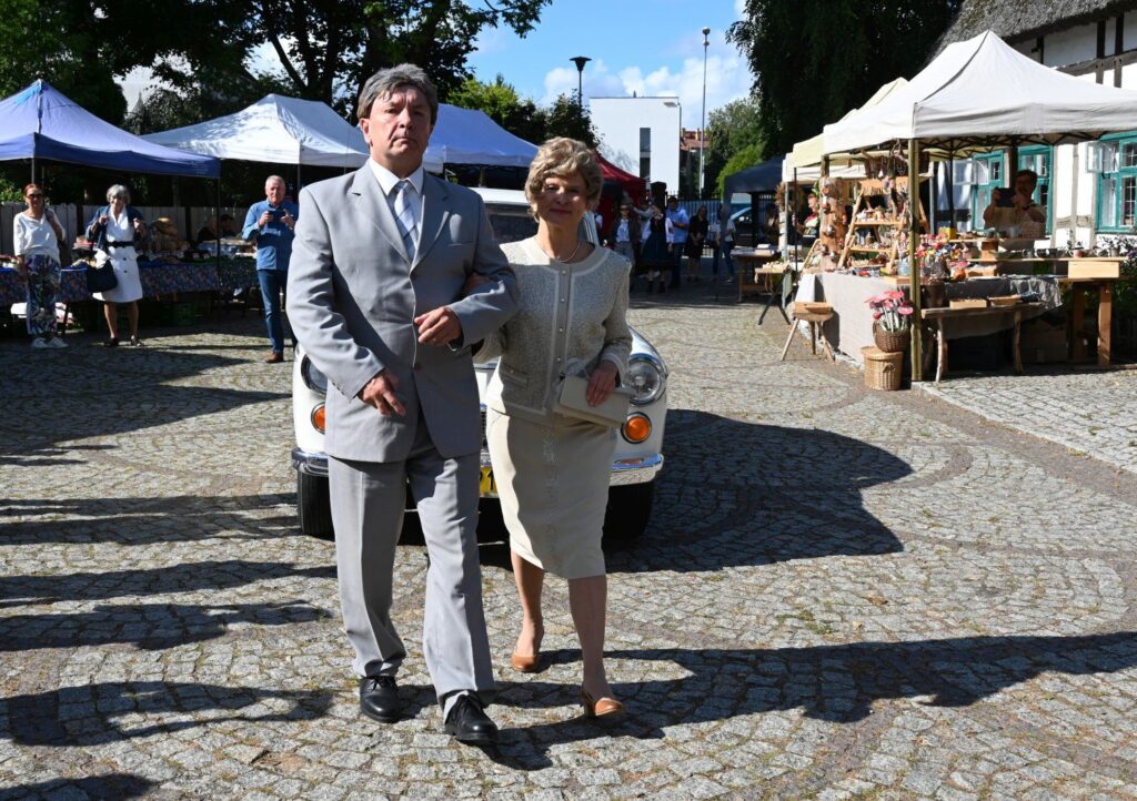Para w eleganckich strojach idąca przed zabytkowym samochodem podczas uroczystości dożynkowych (kliknięcie powiększy zdjęcie).