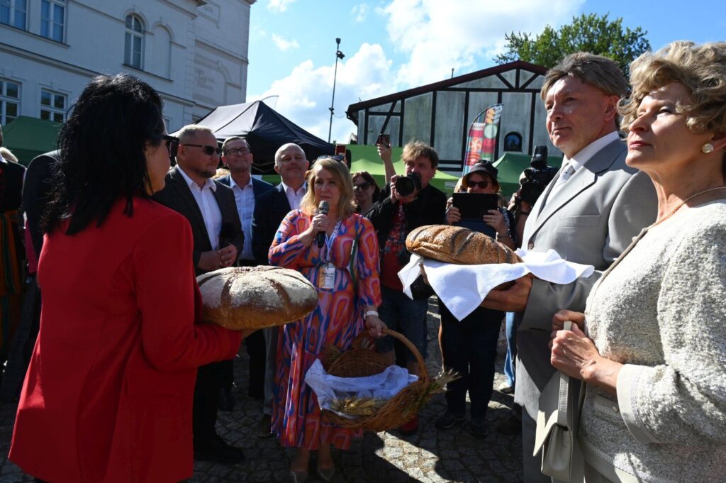 Powitanie gości chlebem podczas festynu dożynkowego, wokół zgromadzeni uczestnicy wydarzenia (kliknięcie powiększy zdjęcie).