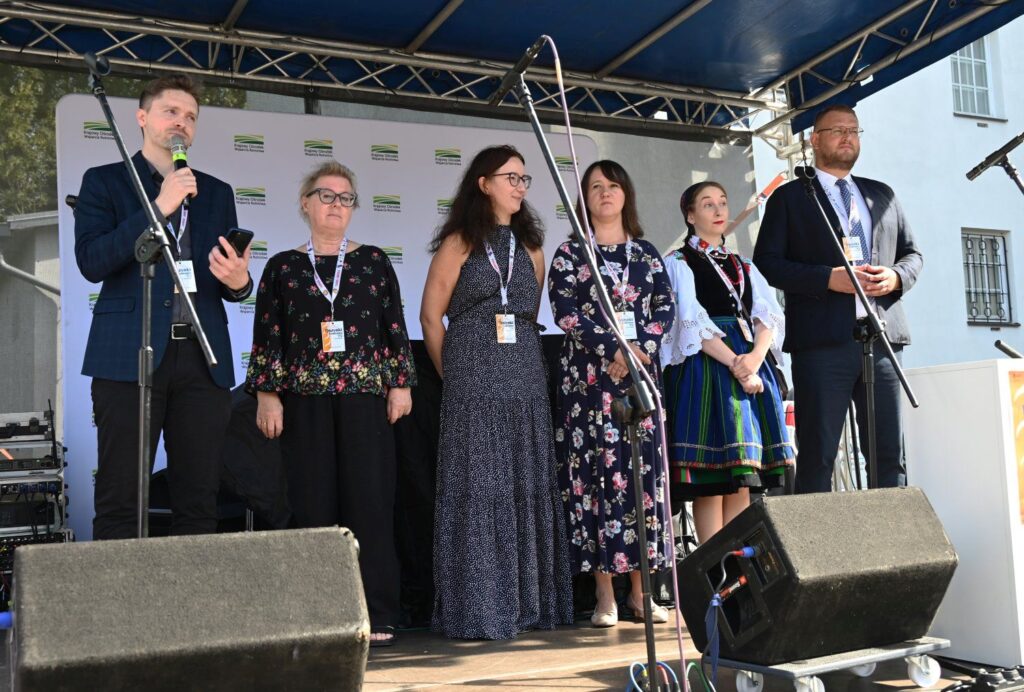 Osoby stojące na scenie podczas oficjalnego otwarcia wydarzenia, jedna z nich przemawia do zgromadzonej publiczności (kliknięcie powiększy zdjęcie).