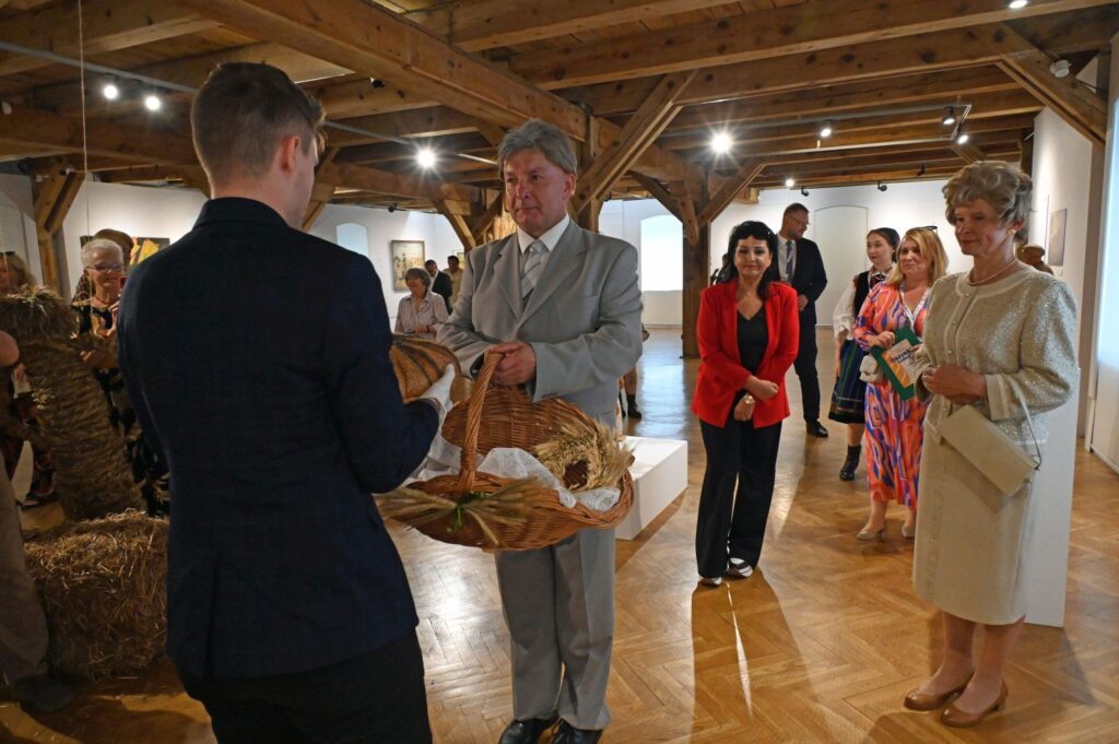 Przekazanie chleba dożynkowego w sali muzealnej, uczestnicy stoją wokół w odświętnych strojach (kliknięcie powiększy zdjęcie).