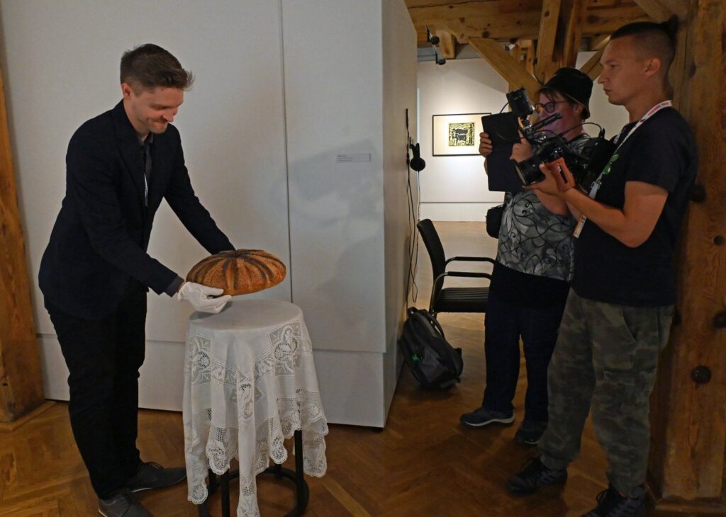 Mężczyzna odkładający bochen chleba na stoliku podczas dokumentowania wydarzenia przez operatora kamery (kliknięcie powiększy zdjęcie).