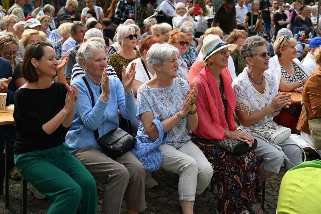 Publiczność siedząca na ławkach i oklaskująca występ artystyczny (kliknięcie powiększy zdjęcie).