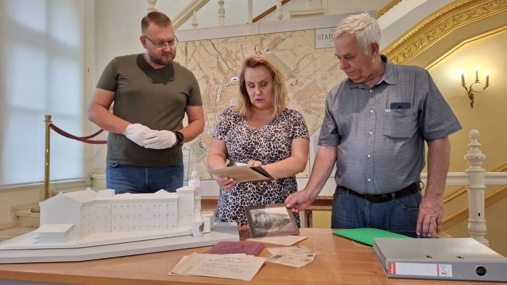 Trzy osoby stoją za stołem, na którym znajdują się archiwalne zdjęcia i dokumenty przekazane do Muzeum. W tle klatka schodowa i wielkoformatowa mapa Koszalina (kliknięcie spowoduje powiększenie zdjęcia).