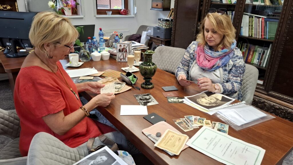 Dwie kobiety siedzą przy stole, w biurze, i przeglądają archiwalne zdjęcia i dokumenty. Stół wypełniony jest innymi artefaktami przekazanymi do muzeum (kliknięcie spowoduje powiększenie zdjęcia).