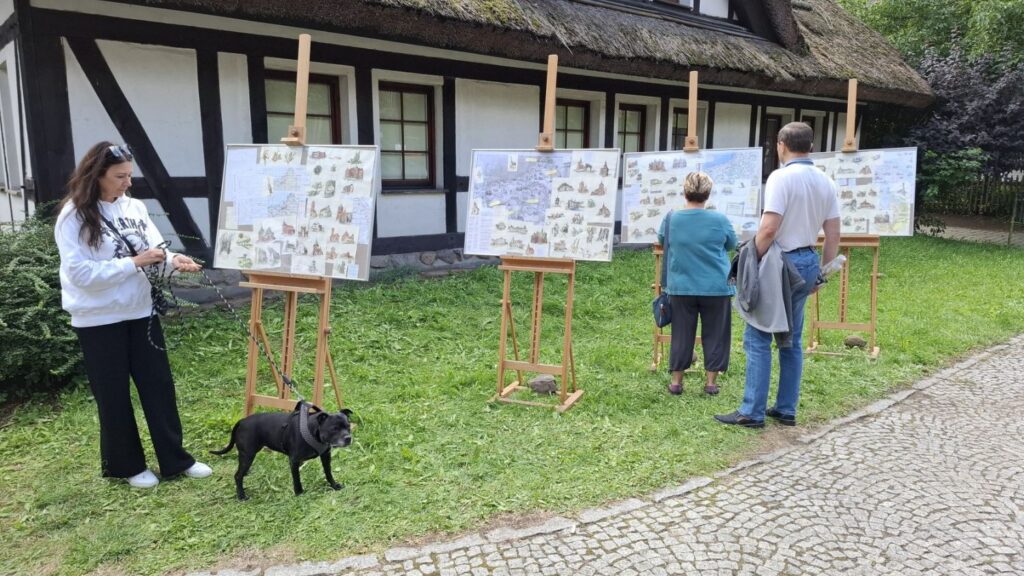 Trzy osoby stoją przed sztalugami z planszami o obiektach architektonicznych i je oglądają. Po lewej stronie kobieta z psem. W tle zabudowania biurowe muzeum (kliknięcie spowoduje powiększenie zdjęcia).