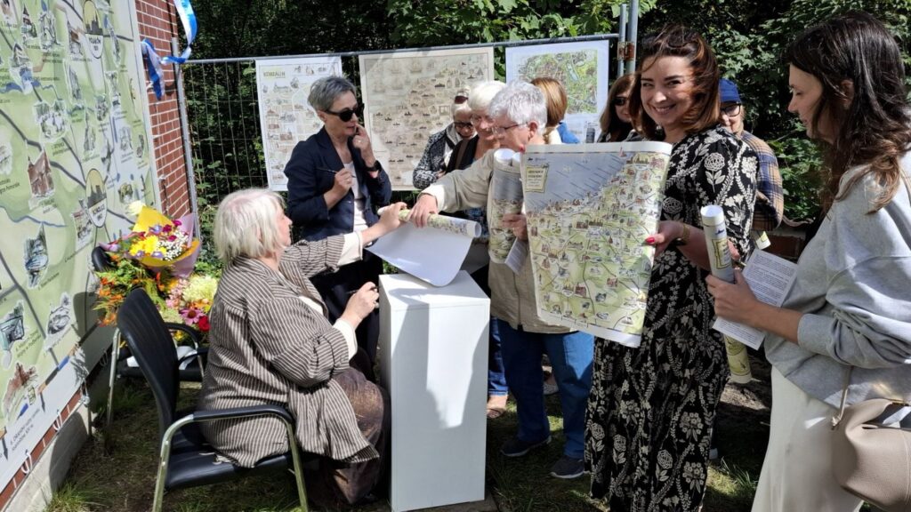 Grupka ludzi stoi w kolejce po autografy autorek mapy "Wędrownik koszaliński". Po lewej widoczna mapa w dużym formacie, a w tle plansze z wydrukami innych map (kliknięcie spowoduje powiększenie zdjęcia).