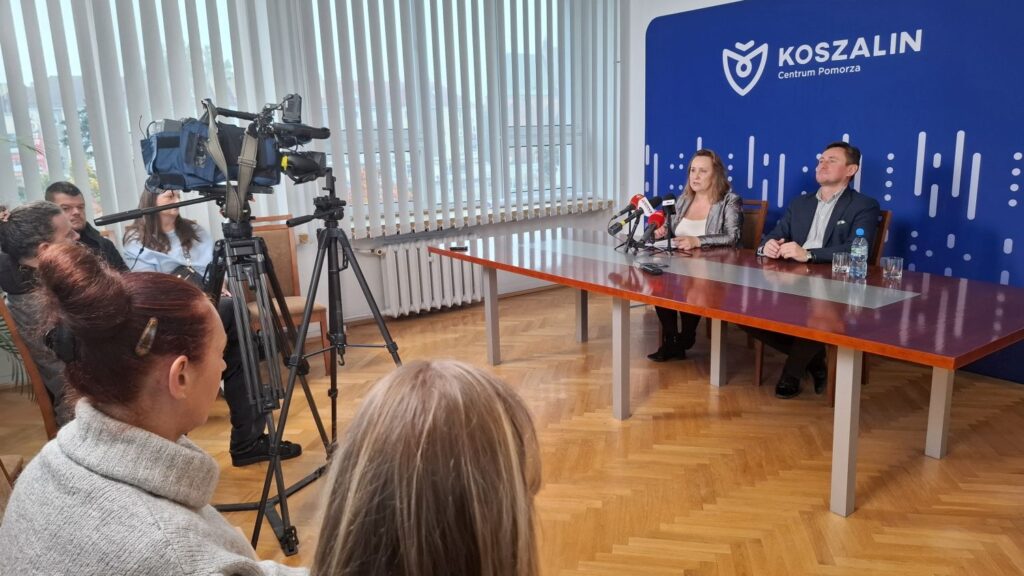 Dwie osoby siedzą przy stole, na którym stoją mikrofony, po lewej - pracownicy mediów i kamera na statywie (kliknięcie spowoduje powiększenie zdjęcia).