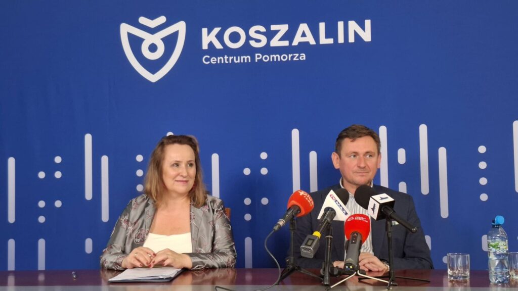 Dwie osoby siedzą przy stole, przed mikrofonami, w tle niebieska plansza z napisem Koszalin Centrum Pomorza (kliknięcie spowoduje powiększenie zdjęcia).