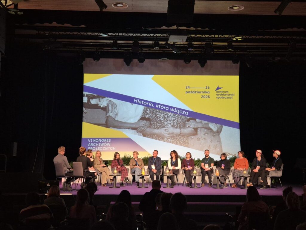 Szerokie ujęcie sceny podczas Kongresu Archiwów Społecznych, na której odbywa się dyskusja panelowa z udziałem wielu prelegentów (kliknięcie powiększy zdjęcie).