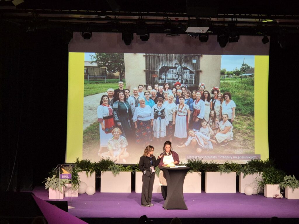 Prezentacja projektu podczas kongresu – prowadzące stoją na scenie przed dużym ekranem z fotografią grupową (kliknięcie powiększy zdjęcie).