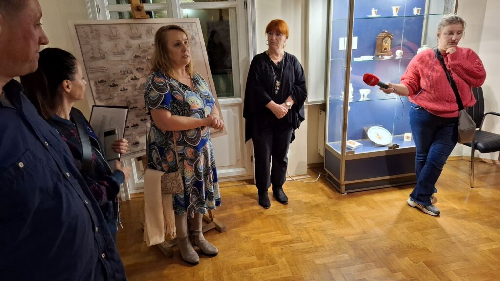 Dyrektor muzeum otwiera spotkanie, a uczestniczy czekają na zwiedzanie, po lewej stoi reporterka z mikrofonem, a za nią przeszklona gablota z artefaktami (kliknięcie powiększy zdjęcie).