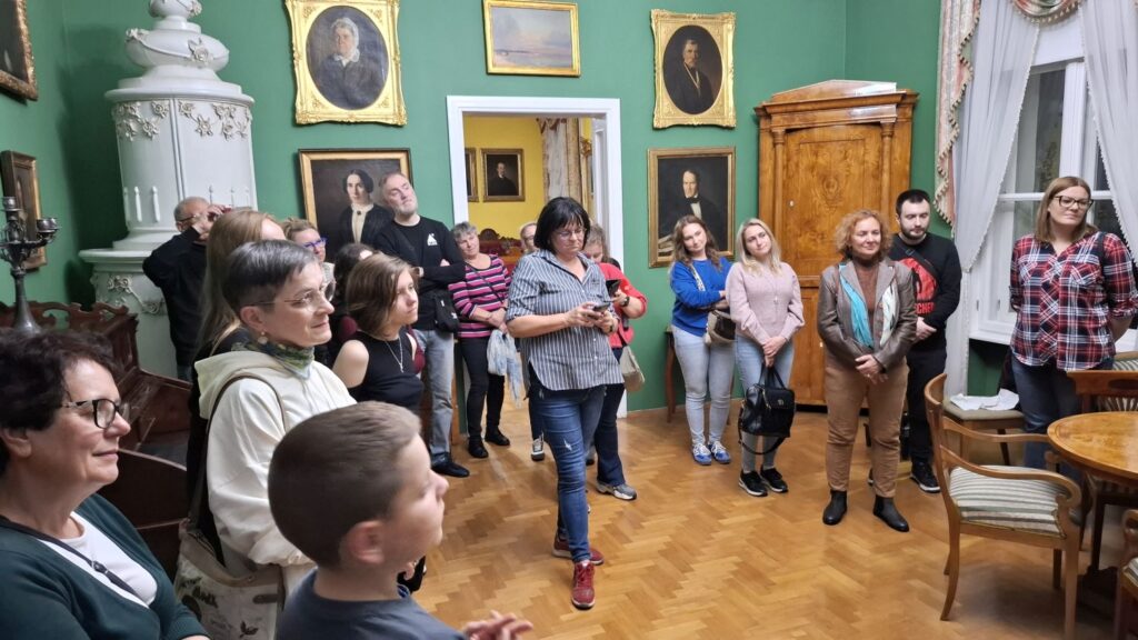 Grupa zwiedzających zgromadzona w zabytkowej sali muzealnej podczas oprowadzania po wystawie, w tle stoją zabytkowe meble a na ścianach wiszą obrazy (kliknięcie powiększy zdjęcie).