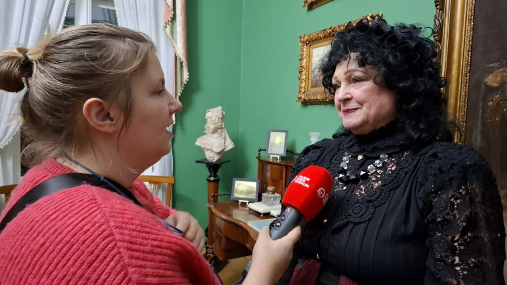 Reporterka radiowa trzyma mikrofon i przeprowadza wywiad z uczestniczką wydarzenia w sali muzealnej podczas (kliknięcie powiększy zdjęcie).