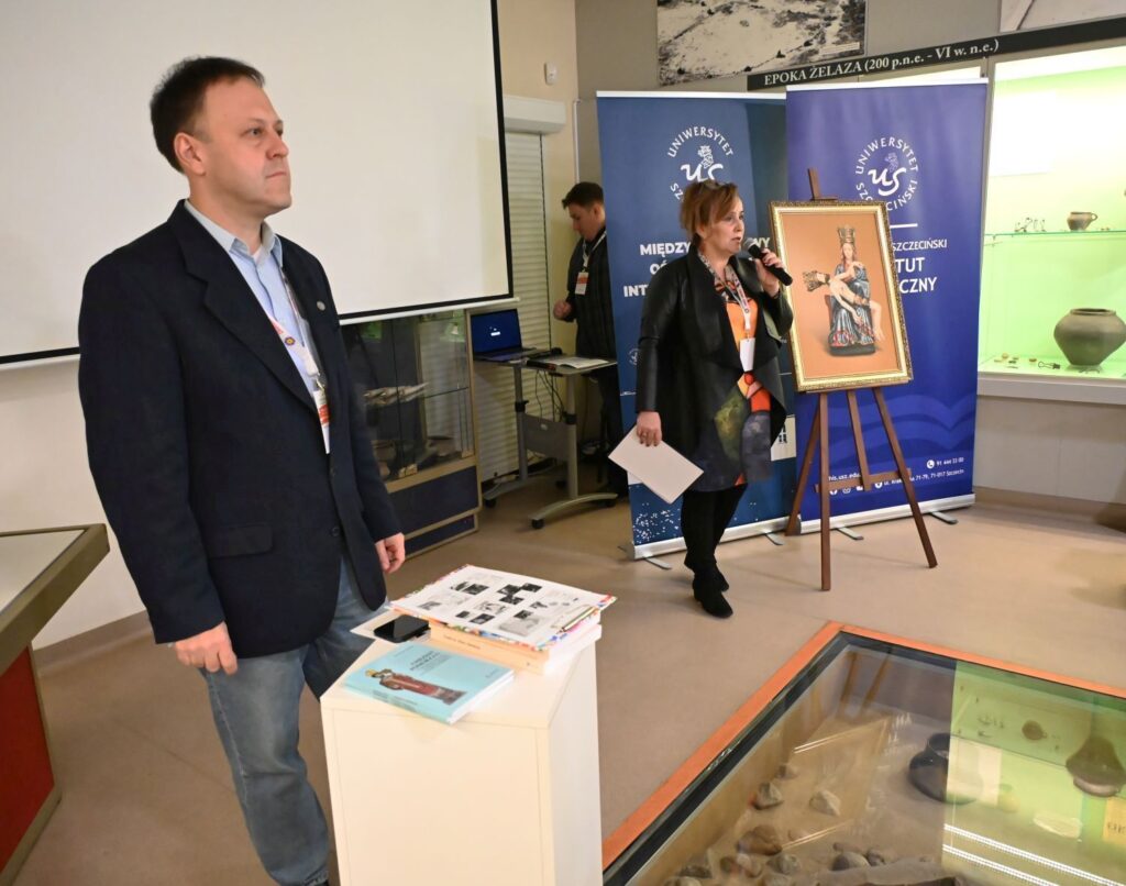 Mężczyzna stoi przy gablocie muzealnej podczas konferencji, w tle dyrektor muzeum rozpoczyna wydarzenie (kliknięcie powiększy zdjęcie).