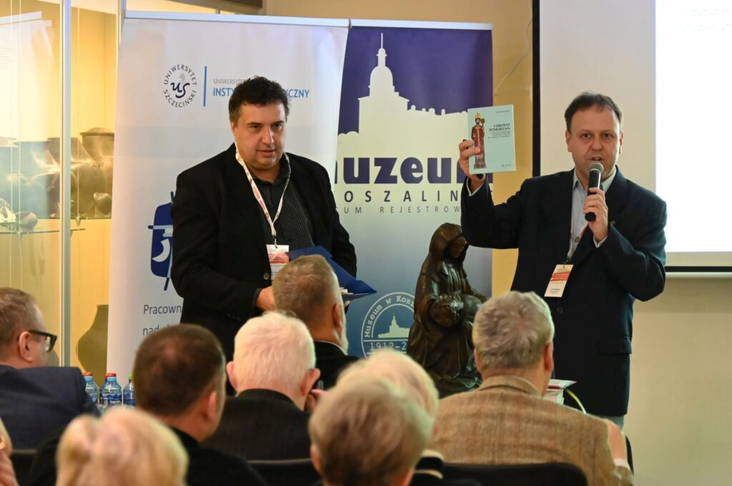 Dwóch mężczyzn na scenie podczas wystąpienia, jeden trzyma publikację, drugi przemawia do publiczności (kliknięcie powiększy zdjęcie).