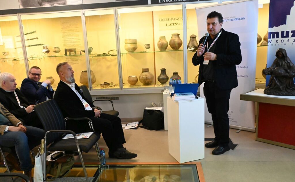 Prelegent przemawia do zgromadzonych uczestników konferencji, siedzących w sali muzealnej, w tle gabloty z eksponatami archeologicznymi (kliknięcie powiększy zdjęcie).