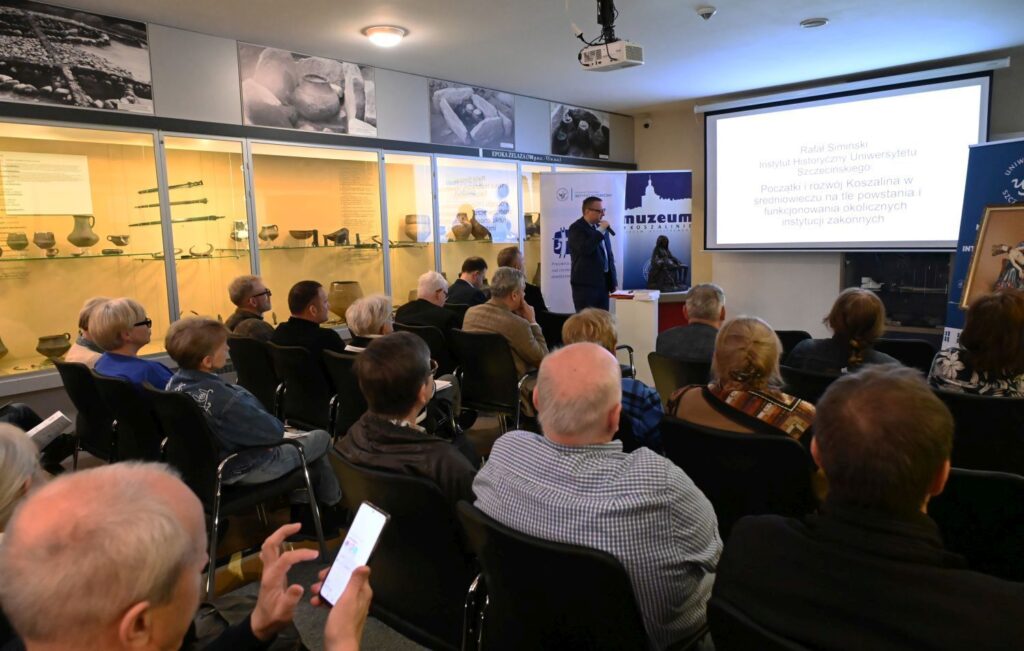 Uczestnicy konferencji oglądają prezentację na ekranie w sali wystawy archeologicznej (kliknięcie powiększy zdjęcie).