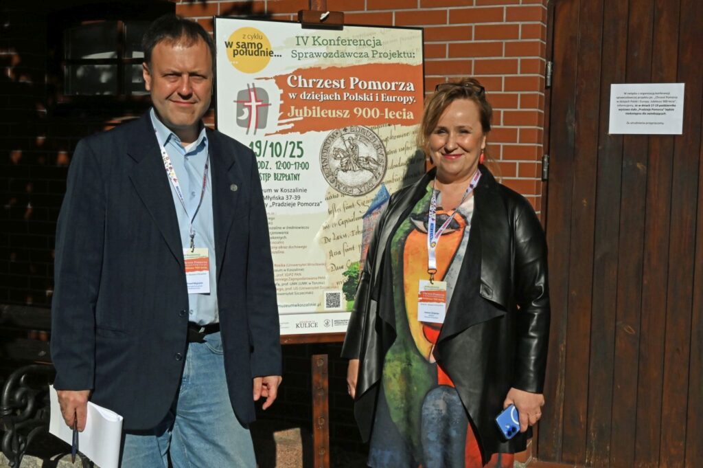 Kobieta i mężczyzna stoją przed plakatem konferencji dotyczącym chrztu Pomorza i jubileuszu 900-lecia, mają identyfikatory na szyjach (kliknięcie powiększy zdjęcie).