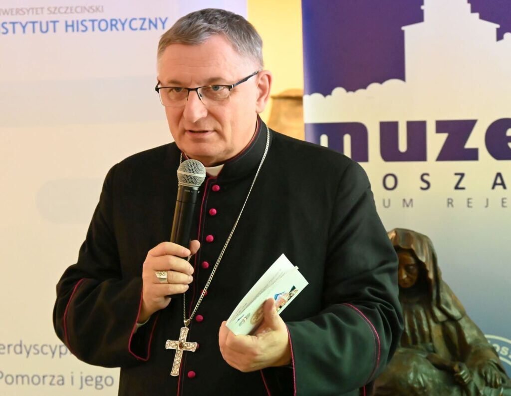 Duchowny przemawia do uczestników konferencji, trzymając mikrofon i publikację (kliknięcie powiększy zdjęcie).