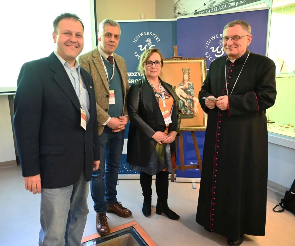 Uczestnicy konferencji pozują do wspólnego zdjęcia przy obrazie na sztaludze (kliknięcie powiększy zdjęcie).