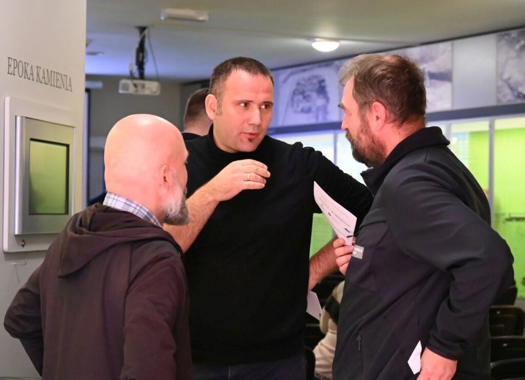 Trzech mężczyzn prowadzi rozmowę w kuluarach konferencji, jeden z nich gestykuluje podczas wypowiedzi (kliknięcie powiększy zdjęcie).